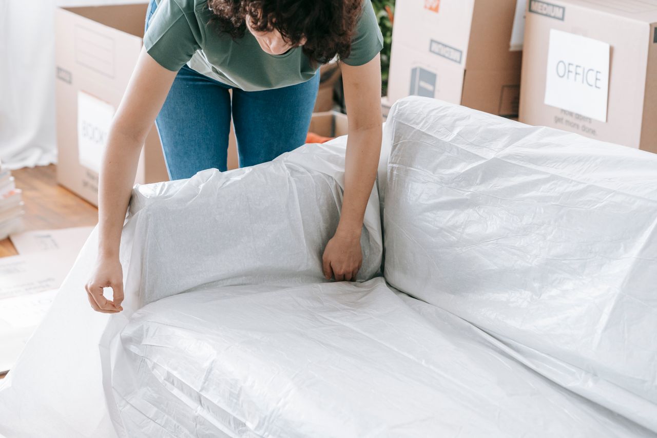 team-03 A woman carefully wraps a sofa in protective material while packing boxes in a bright room.